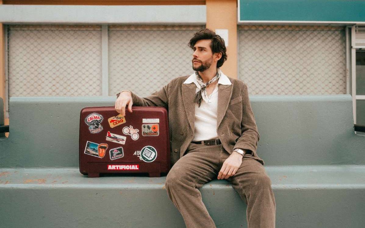 Musiker Alvaro Soler sitzt in einem Pressefoto von 2025 auf einer Bank neben einem Koffer